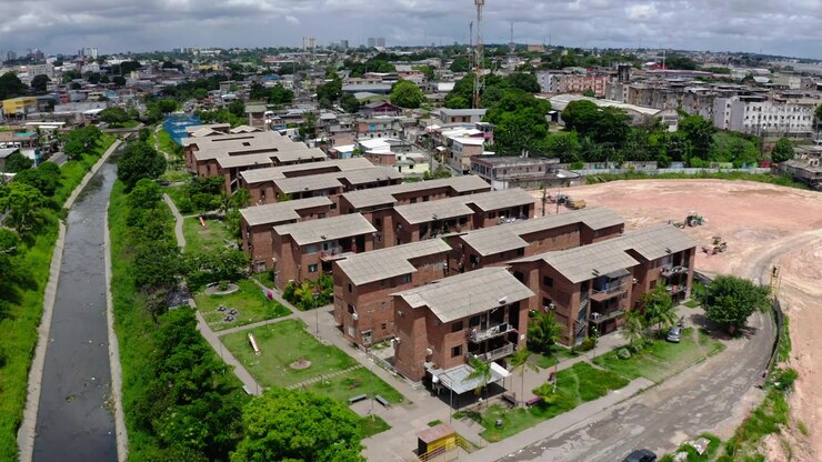 Drone aereo che si allontana lentamente sopra file di blocchi di edilizia popolare in mattoni di un conjunto habitacional vicino all'igarape canalizzato a Manaus, parchi giochi, palme, lotto livellato, skyline, soleggiato, Brasile