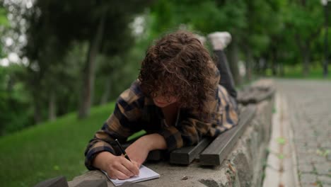 Happy-brunette-girl-with-curly-hair-makes-notes-in-her-notebook-while-lying-on-a-bench-in-the-park