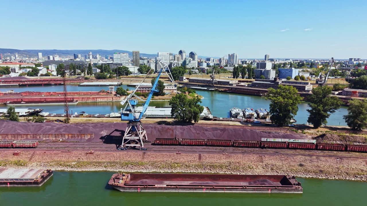 Area industriale con gru, barche e vagoni ferroviari vicino a un fiume in una città di giorno.