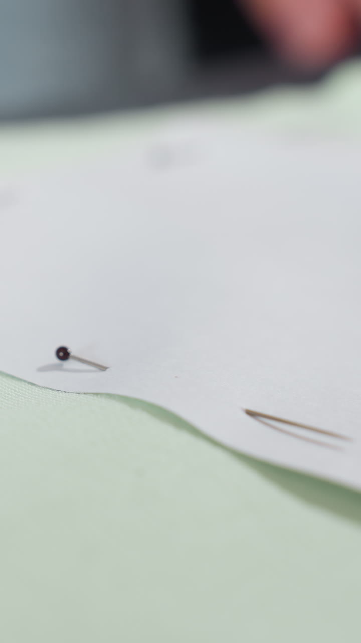 closeup fingers pinning paper pattern on green cutting mat, slow careful placement of black head pins along paper edge, shallow depth of field, quiet studio light, meticulous craft preparation