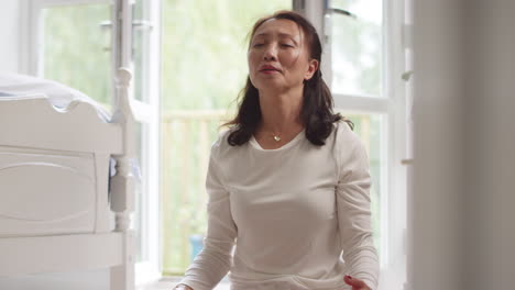 Mature-Asian-woman-in-pyjamas-sitting-on-bedroom-floor-meditating-in-yoga-pose---shot-in-slow-motion