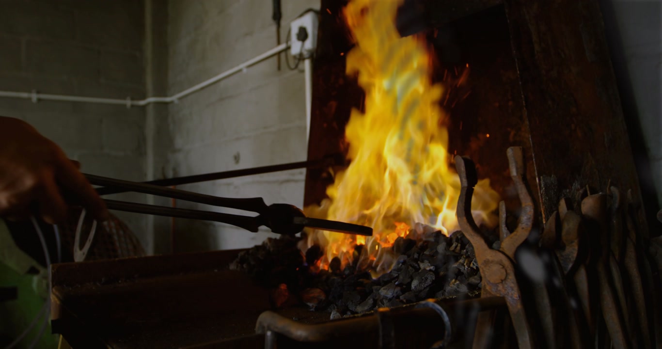 Metalworker heating metal in coal fire with tongs, sparking as grill casting dot shadows across arm