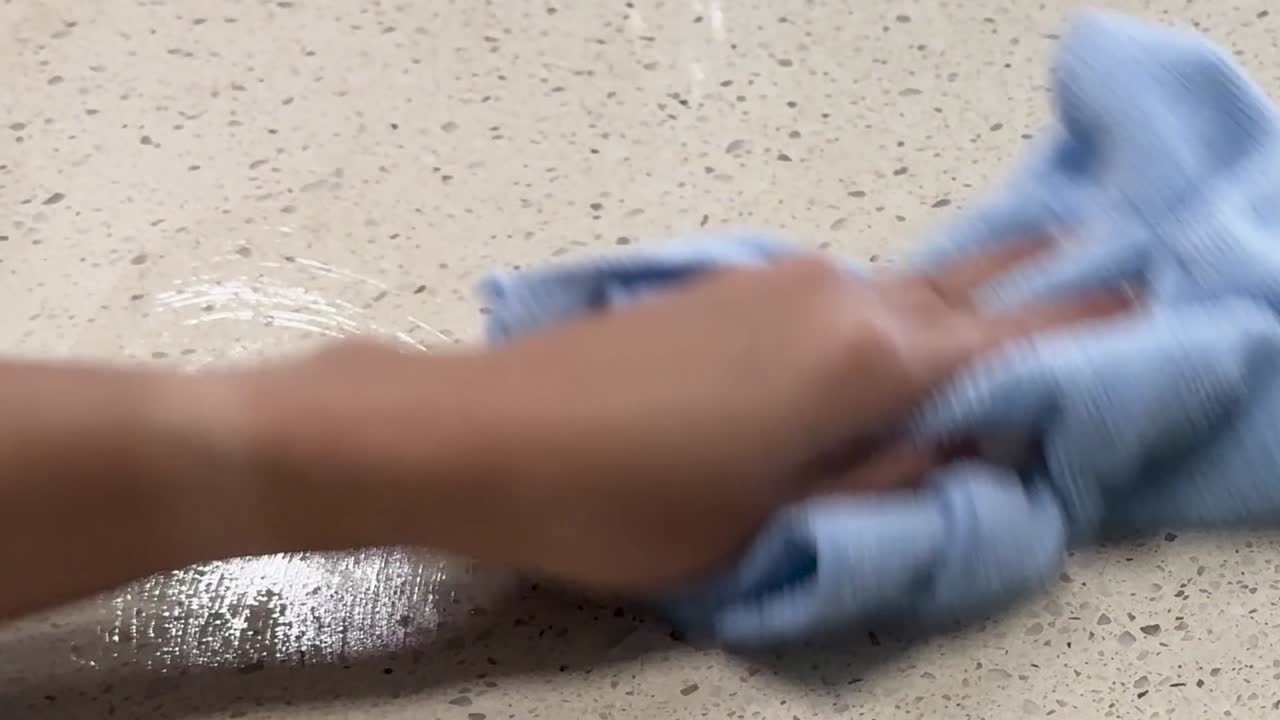 A person sprays cleaner and wipes a coffee stain from a light granite surface