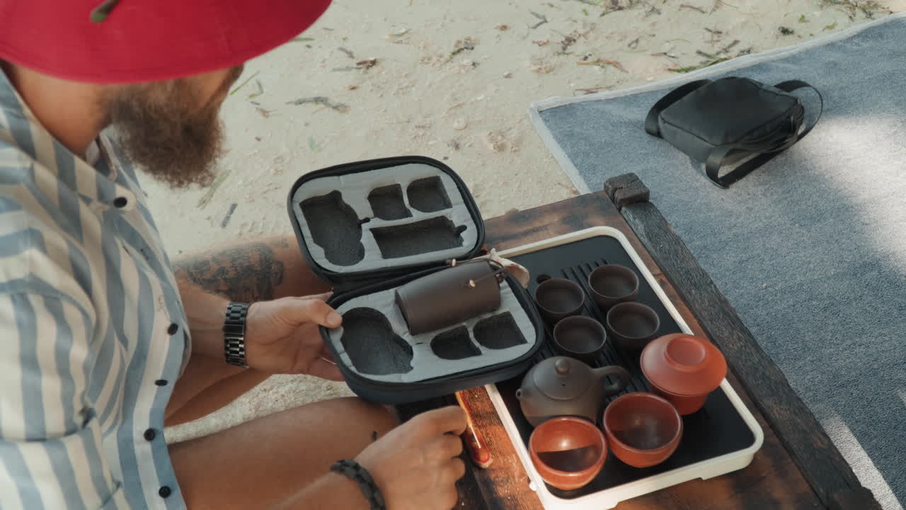 Strandte-kit, kaukasisk man packar upp väska som avslöjar lertekanor och koppar, händer organiserar fack på matta, ovanifrån, solbelysta texturer, produktlivsstil, noggrant hantverksfokus.