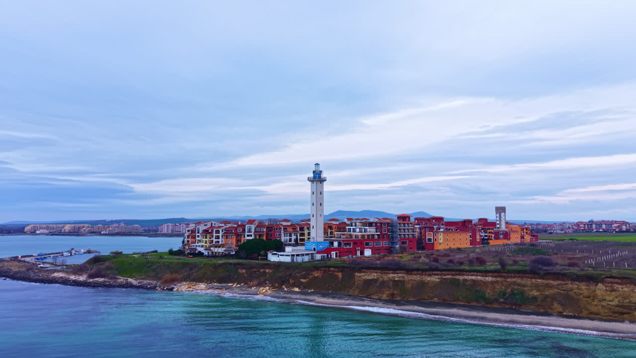 Vista marittima di un faro vicino a una città costiera con edifici sulla riva e acqua in primo piano sotto un cielo nuvoloso