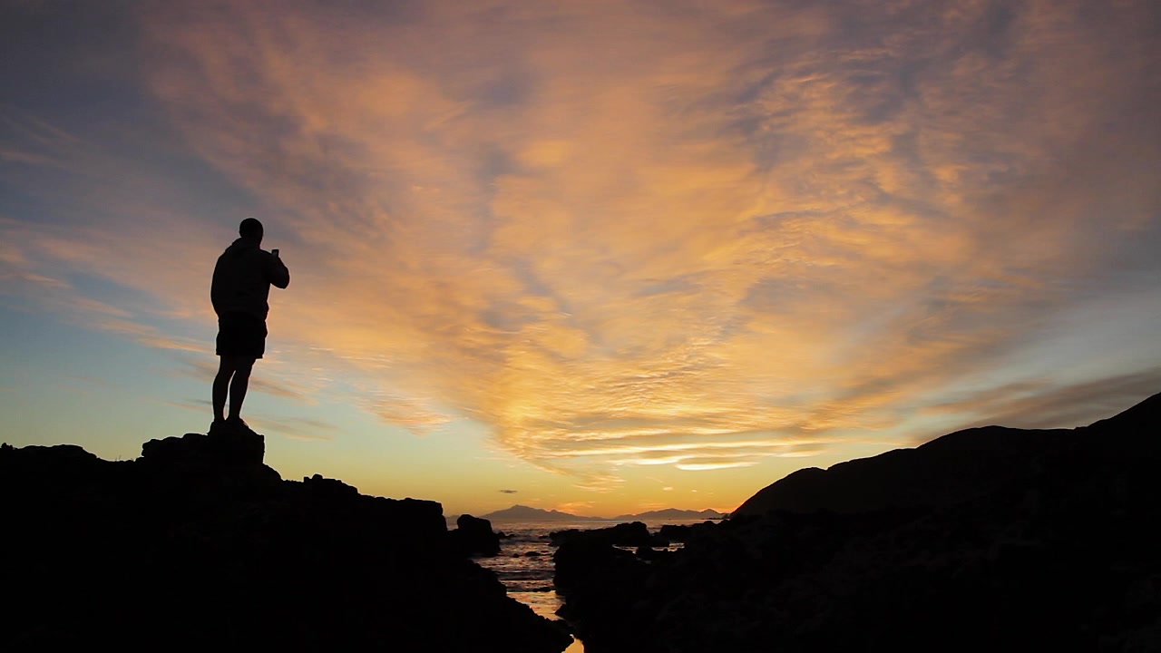 The silhouette of a person looking at the sun set and taking a photo with their phone.