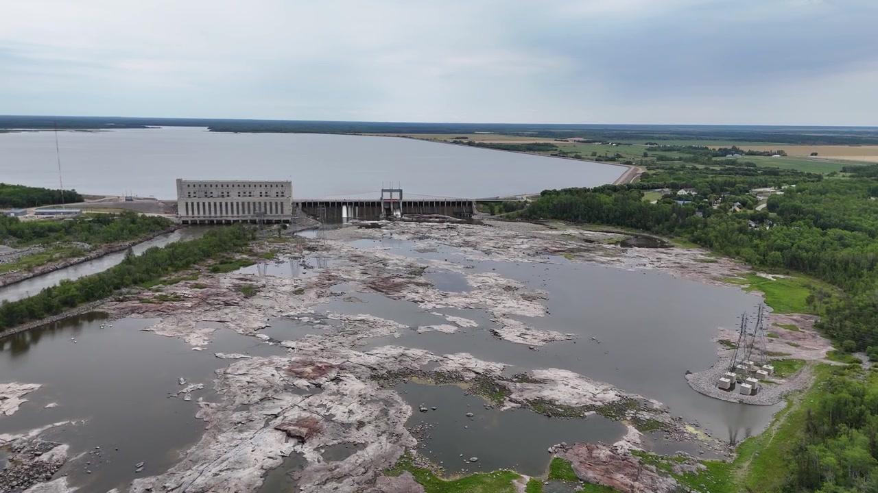 Ampia ripresa aerea con drone in volo in avanti da lontano verso la Centrale elettrica di Seven Sisters, che mostra rocce esposte dello Scudo Canadese, canale di sfioro, lago artificiale dietro la diga.