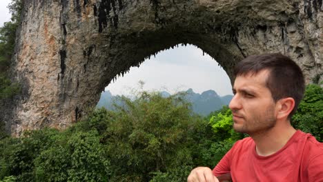 hombre sonríe a la cámara con el arco natural de la colina de la luna y la vegetación exuberante en el fondo en yangshuo, china