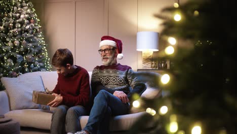 happy cute teen boy giving xmas present to happy old grandpa while sitting at cozy decorated home near new year tree