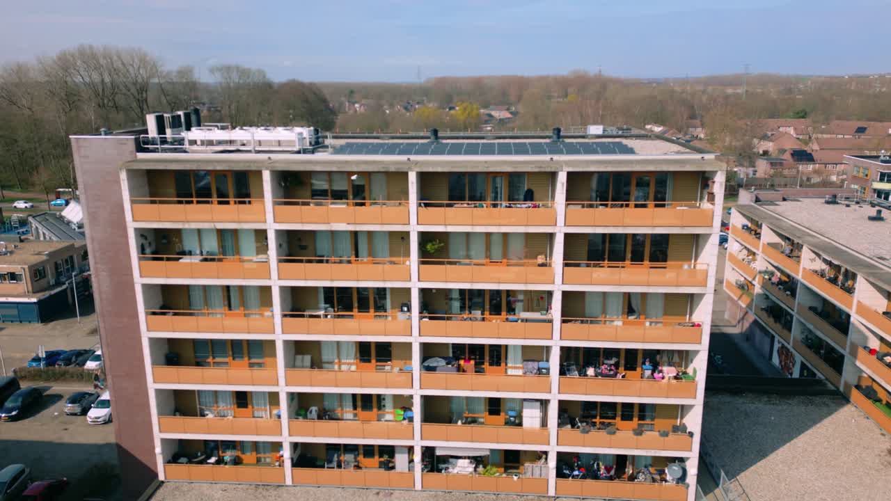 Detaljeret luftfoto af en moderne lejlighedsbygning med mange balkoner på hver etage. Taget er synligt med installerede solpaneler, der fremhæver indsatsen for bæredygtig energi. Biler parkeret på.