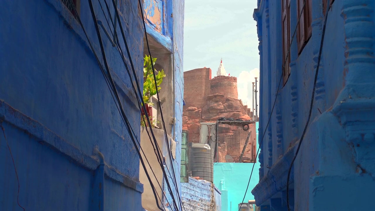Inquadratura statica a media distanza che incornicia le maestose mura in arenaria rossa del Forte di Mehrangarh tra gli stretti edifici del vicolo blu a Jodhpur, Rajasthan, India