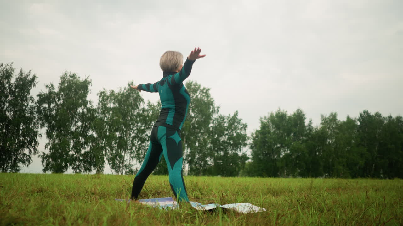 mujer de traje negro verde en una alfombra de yoga practicando yoga en una postura triangular hacia adelante en un campo abierto de hierba, brazos extendidos, con vegetación y árboles en el fondo bajo un cielo nublado