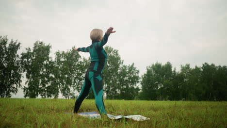 woman in green black suit on yoga mat practicing yoga triangle forward pose in open grassy field, arms extended, with greenery and trees in the background under cloudy skies