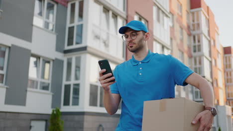 a male mail carrier with round glasses carries boxes for delivery to customers and looks at his mobile phone. search through the mobile phone for the delivery address. payment terminal.