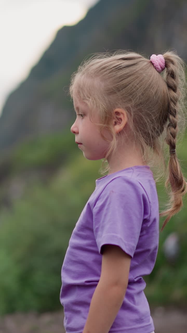 Calm little child in pastel t-shirt stands on hill looking at large distant mountain silhouette on nasty day close side view slow motion