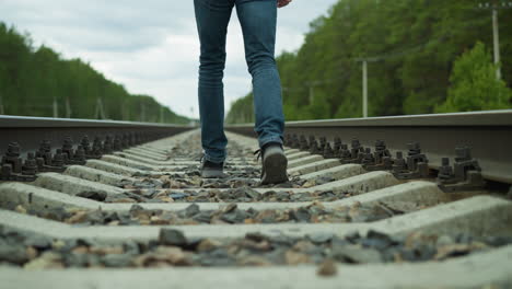 a close-up view of a man's legs, dressed in jeans and canvas shoes, walking alone on railway tracks surrounded by dense trees and electric poles