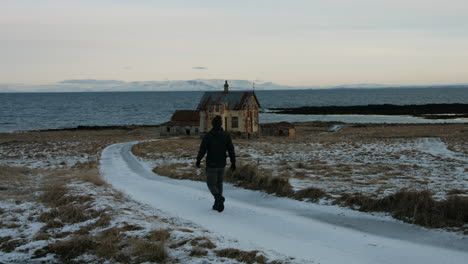 joven solitario caminando por un camino de campo curvo, casa islandesa abandonada en el frío profundo - tiro constante