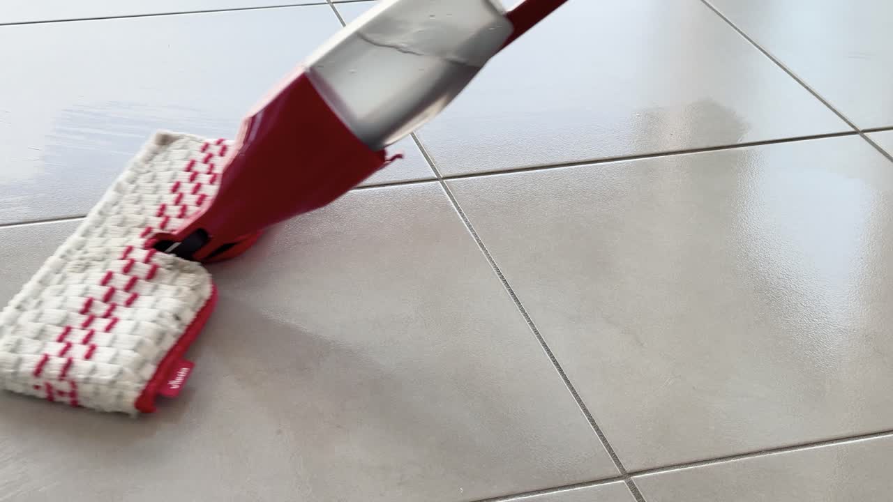 A red spray mop glides over glossy kitchen floor tiles, cleaning water with smooth, steady movements in bright natural daylight
