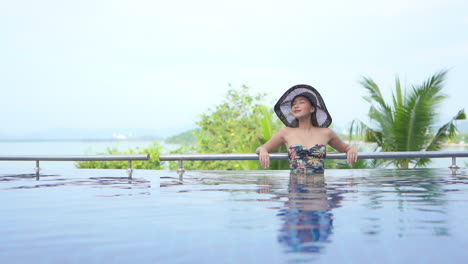 Elegante-Joven-Mujer-Asiática-De-Pie-Junto-Al-Borde-De-La-Piscina-En-Traje-De-Baño-Y-Sombrero