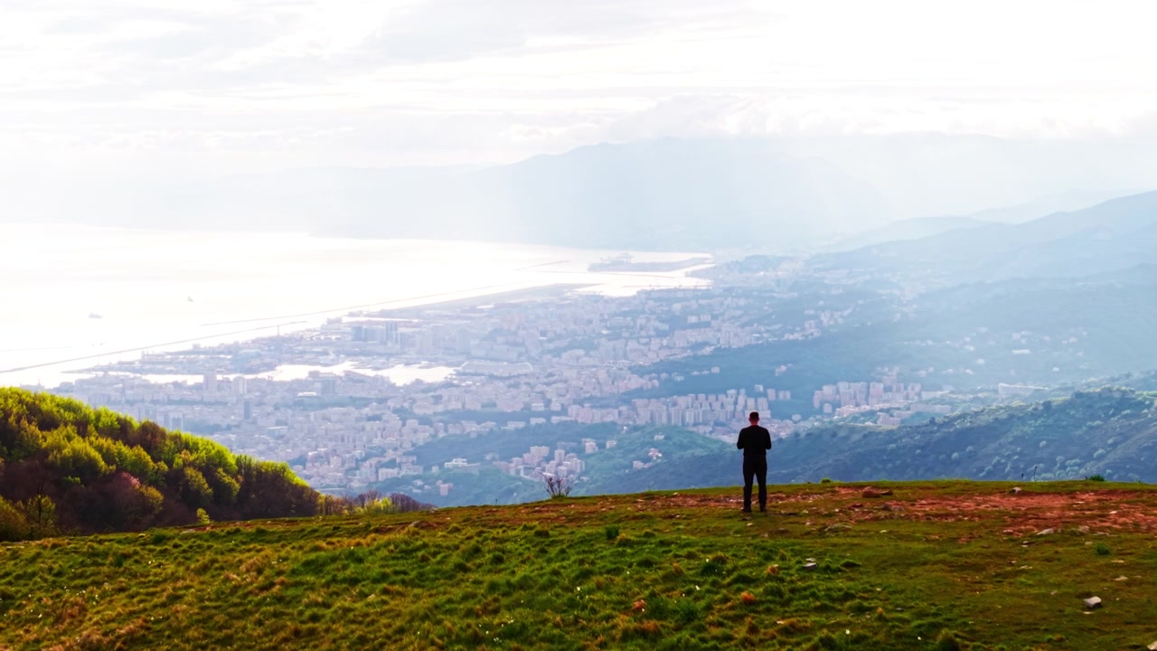 A stunning aerial drone perspective of a filmmaker operating a drone at a mountain summit, capturing the sweeping green valleys of Liguria, Italy.