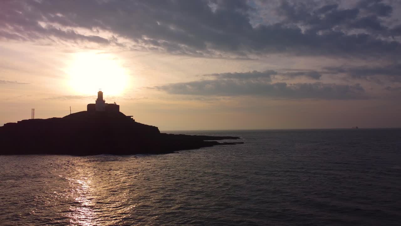 Tomada aérea em avanço lento do Farol de Mumbles ao nascer do sol. Reflexo dourado do sol no oceano com nuvens dinâmicas. Perfeito para turismo, documentários de viagem. País de Gales, Reino Unido. 4K.