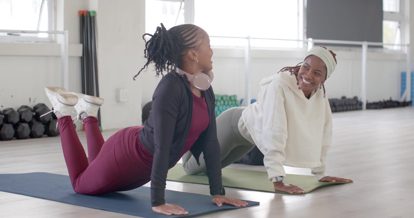 Two adult African women arriving on mats in studio, kneeling, sliding, checking watch, warming up
