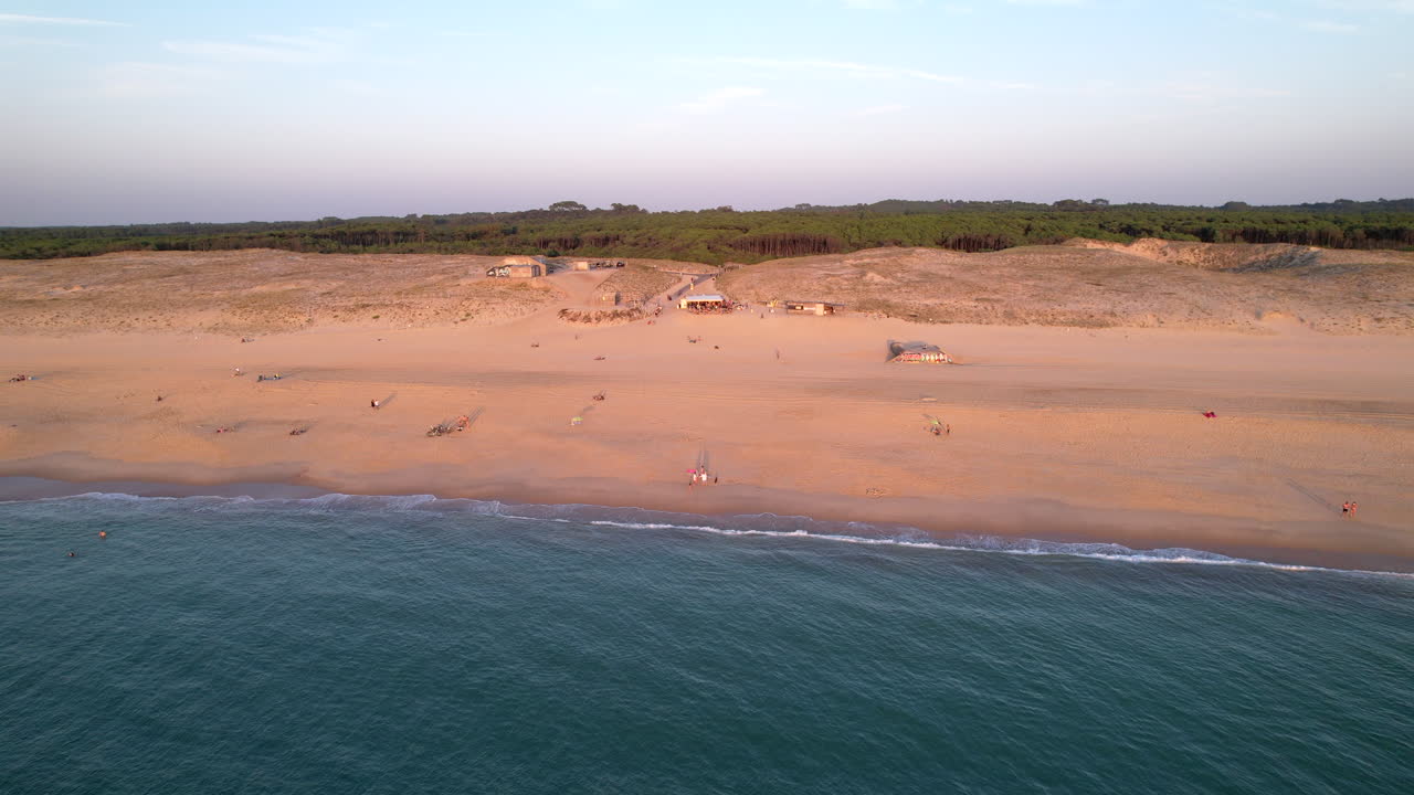 A lateral drone shot showcasing the beach with a few people, water filling the bottom third, and a view of the forest behind, captured on a warm summer evening.
