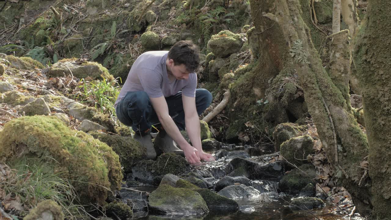 Young man washes face and drinks from woodland stream, brook, creek