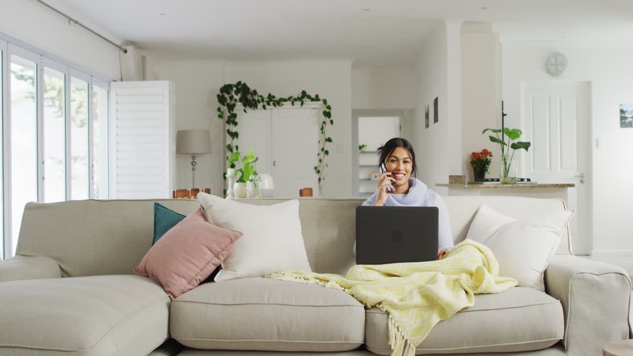 소파에 앉아 스마트폰과 노트북을 사용하는 히자브를 입은 행복한 두 인종 여성의 비디오