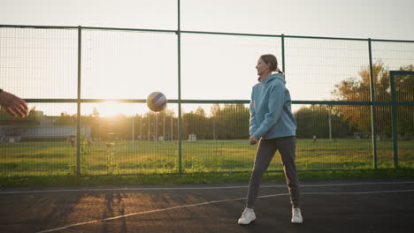 lady in blue hoodie engages in volleyball training session in outdoor court, she hits ball inaccurately, and another lady in green leggings rushes after the ball during the activity at golden hour