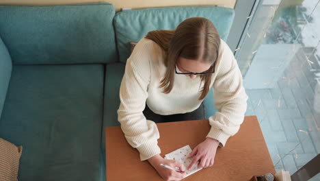 overhead view of young lady in white sweater sketching on paper at wooden table beside window, city pavement and tall buildings faintly visible in soft daylight through glas