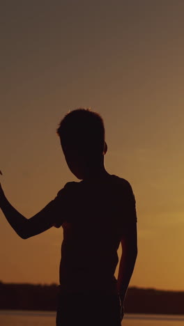 silhouette of little boy sitting alone at the river on sunset background. happy child playing with paper airplane. vertical video