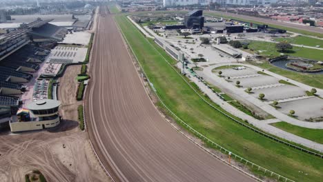 Video-De-Drones-De-Una-Pista-De-Carreras-De-Tierra-De-Caballos,-Con-Un-área-De-Césped-Verde-En-El-Medio-También-Con-Edificios