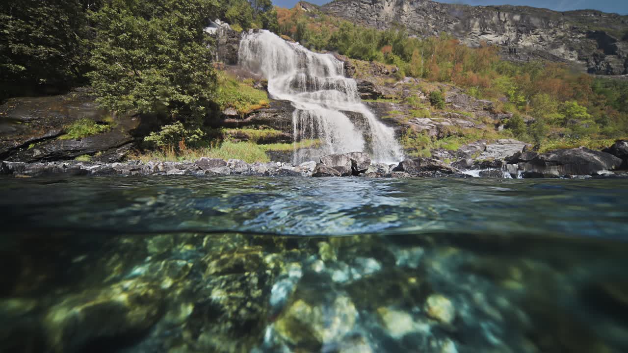 A split over-under view of the waterfall cascading into the fjord. Sun penetrates the water and dances on the rocky bottom.