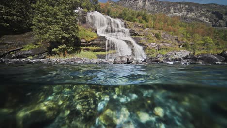 a split over-under view of the waterfall cascading into the fjord. sun penetrates the water and dances on the rocky bottom.