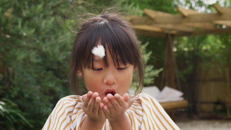 Ritratto di bambina asiatica sorridente con i denti davanti mancanti che si diverte a soffiare una piuma nel giardino di casa.