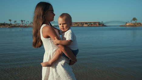 Langes-Haar,-Teenager-Mädchen,-Das-Mit-Einem-Kleinen-Jungen-Auf-Den-Händen-Am-Sonnigen-Strand-Entlang-Spaziert.