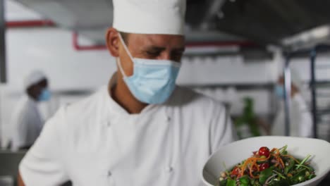 Portrait-of-mixed-race-male-chef-wearing-face-mask-presenting-dish