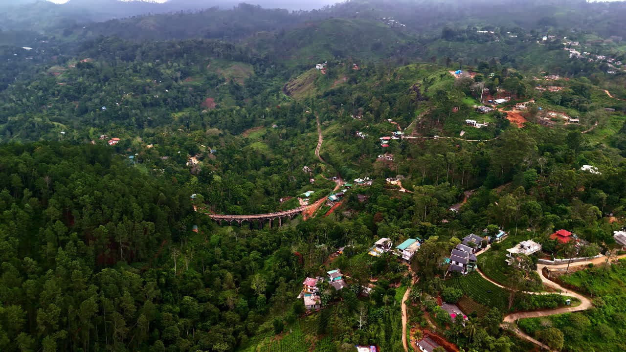 High drone shot over Sri Lanka’s lush Ella hills, with the Nine Arches railway bridge curving through dense forest and scattered village homes in soft mist