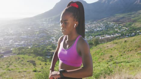 African-american-woman-exercising-outdoors-wearing-wireless-earphones-using-watch-in-countryside