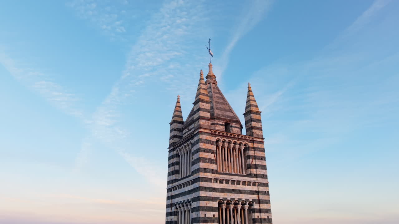 Naher Drohnenflug um die Spitze des Glockenturms des Doms von Siena, dessen Kreuz sich vor dem Sonnenaufgangshimmel abzeichnet.