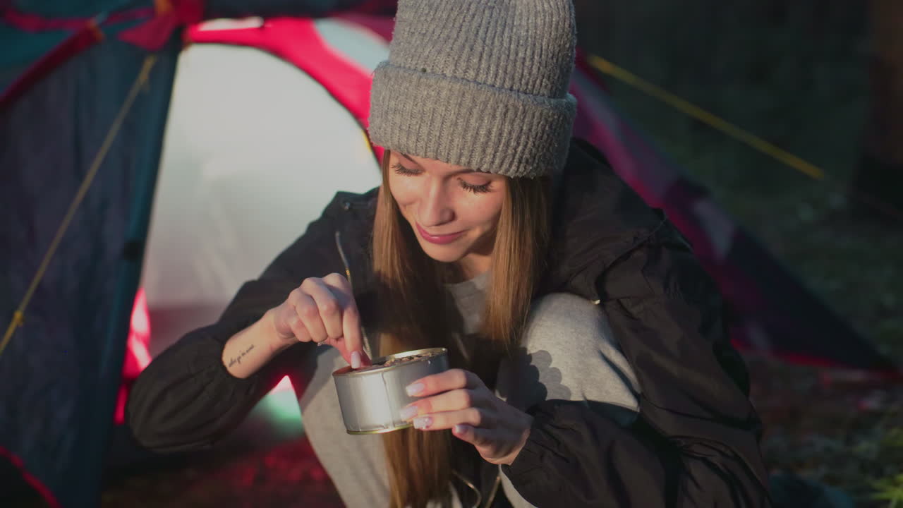 close up of lady squatting outdoors testing can food with warm smile near camp tent during sunset in forest surrounded by grass casual clothing and beanie warm