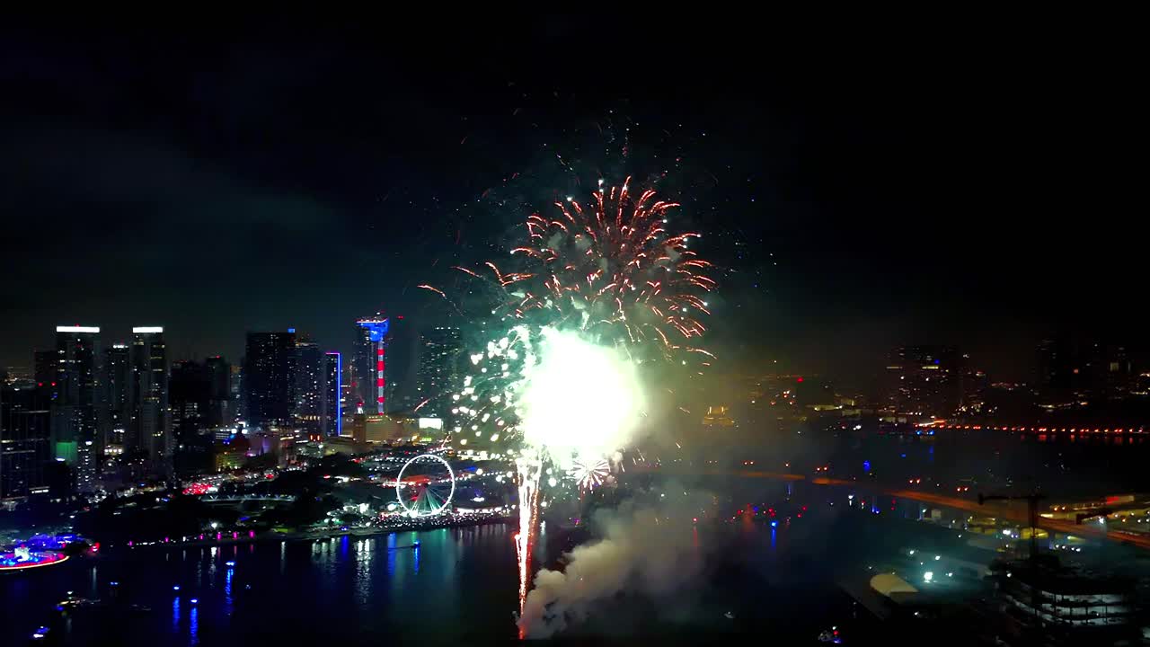 Drone footage soaring high, capturing vivid firework bursts reflecting across the water with glowing lights framing the background
