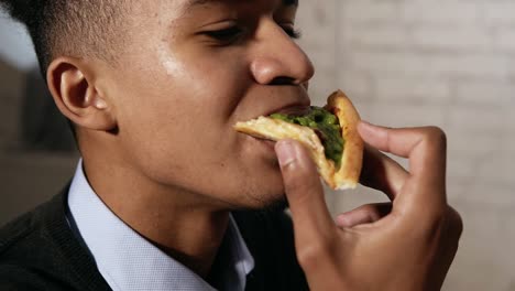 vista de cerca de un joven afroamericano disfrutando de pizza durante el almuerzo en el trabajo. está mordiendo una rebanada en cámara lenta