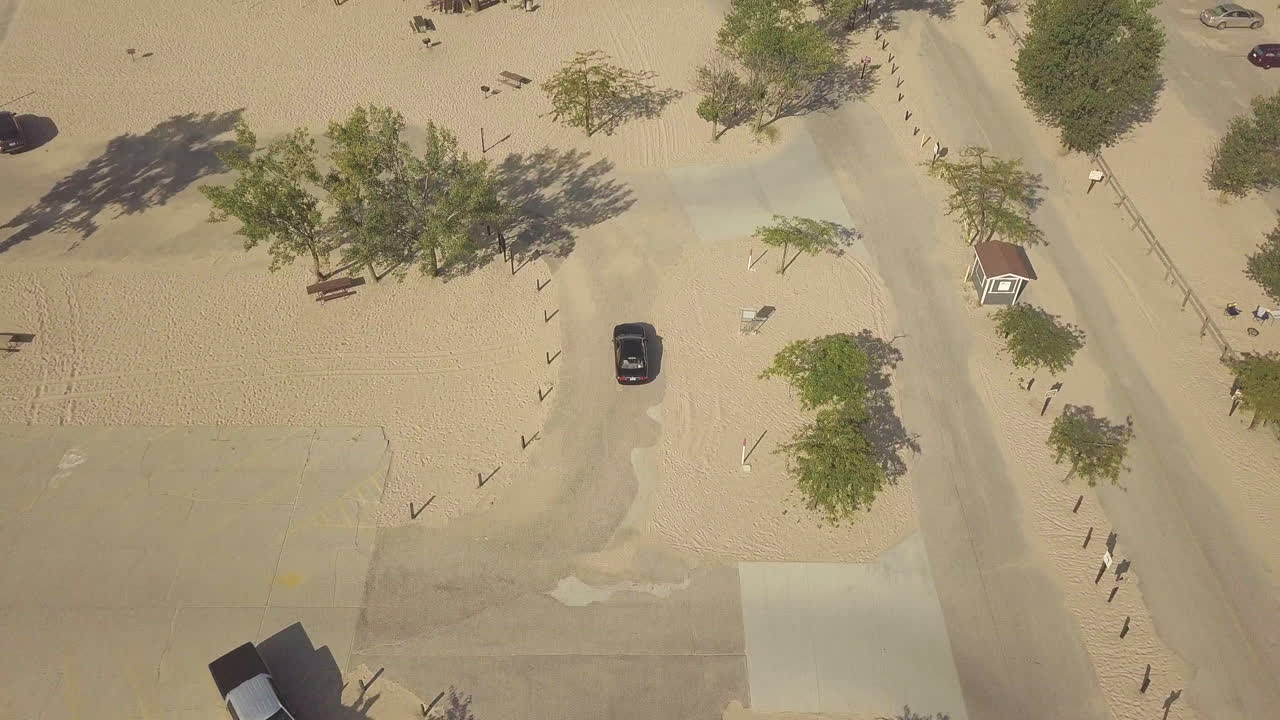 Aerial view of a black car driving through the parking lot at a sandy beach campground.