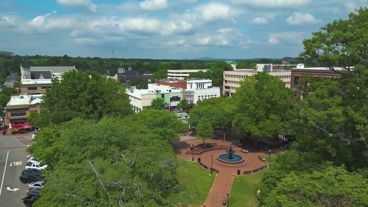 Fountain in Marietta Square Park of american town during sunny and cloudy spring Day. Parking cars in street. Court House buildings in distance. Aerial orbit wide shot.