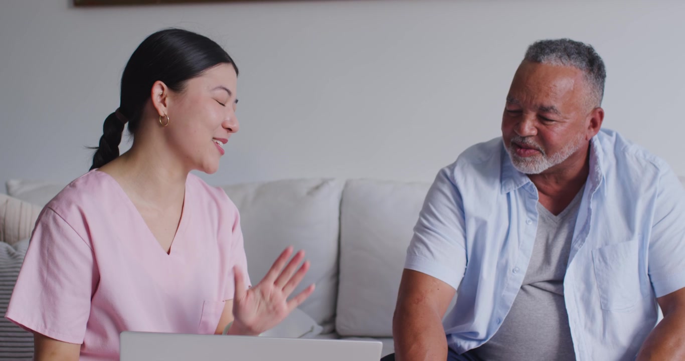 Caregiver wearing scrubs and mature client smiling showing laptop explaining health info on sofa