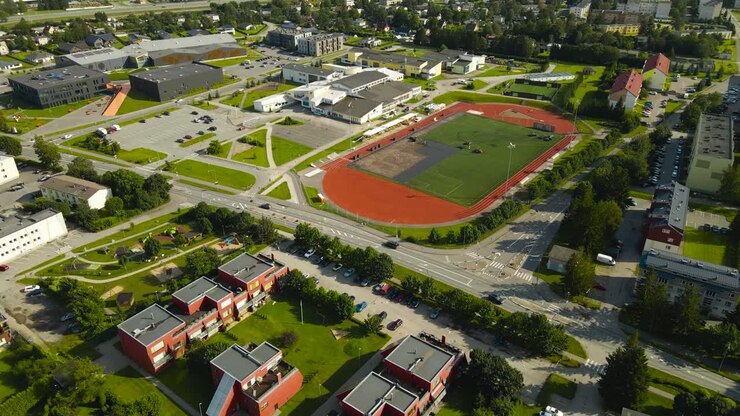 Filmagem aérea de drone orbitando e voando em frente a um grande campo de futebol ou estádio nas cores verde e vermelho que está em construção ou sendo renovado, com trabalhadores removendo a grama verde. Ensolarado.