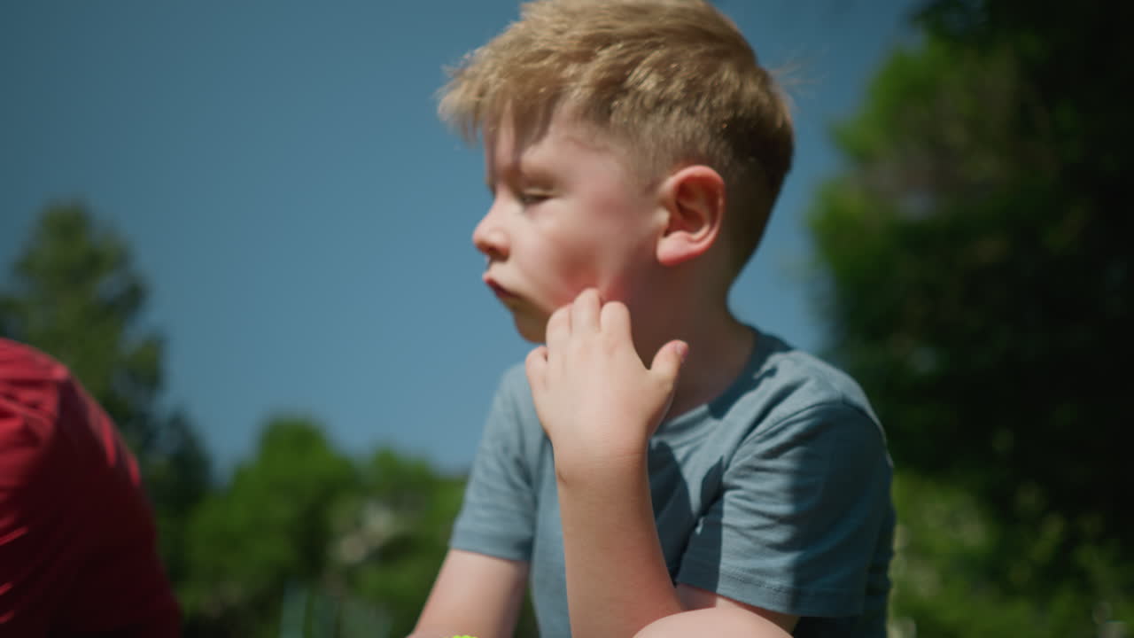 파란 셔츠를 입은 작은 소년은 왼손으로 얼굴을 문지르고, 잔디에 앉아서 가까운 사람을 부분적으로 볼 수 있습니다.