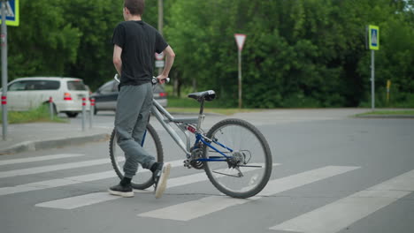 young boy in grey top and black trousers walks alongside his bicycle on a pedestrian crossing, cars are visible, with one in motion and another waiting at the traffic light